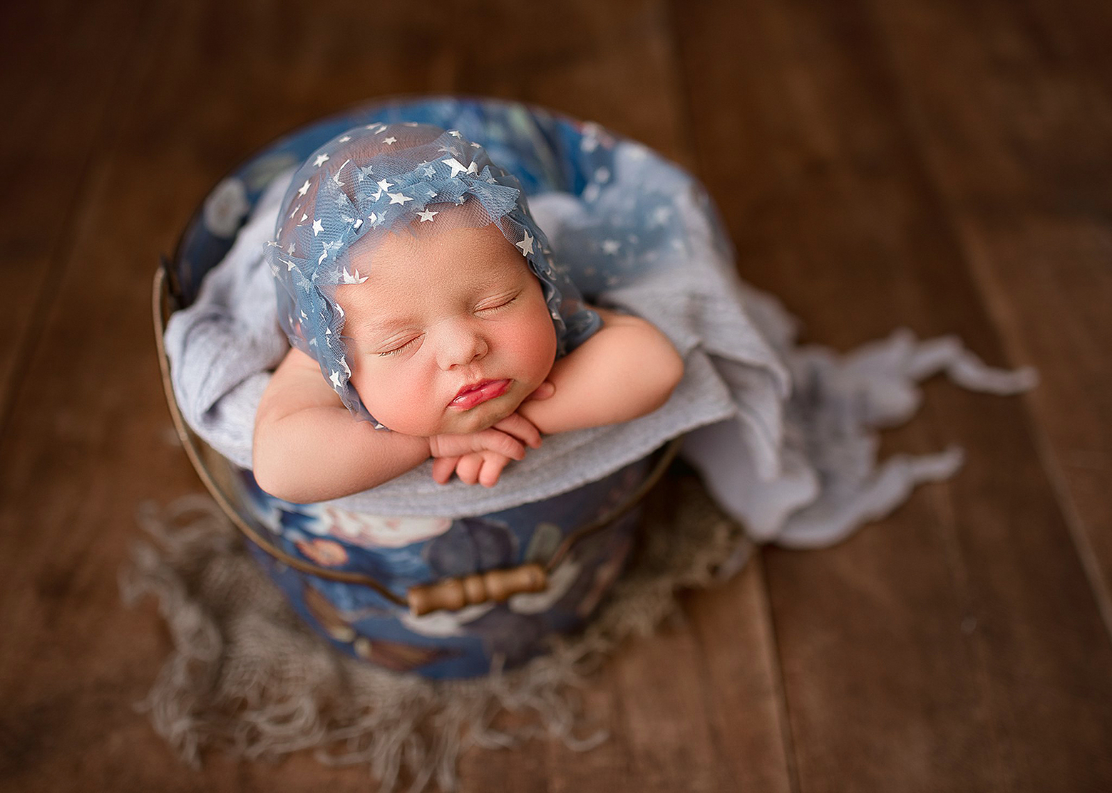 Newborn baby girl sleeping peacefully, wearing a blue bonnet with stars on, photographed in a blue floral bucket